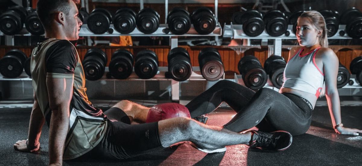A man and woman resting together on the gym floor, surrounded by dumbbells.
