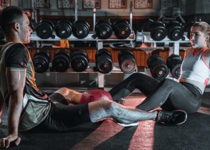 A man and woman resting together on the gym floor, surrounded by dumbbells.